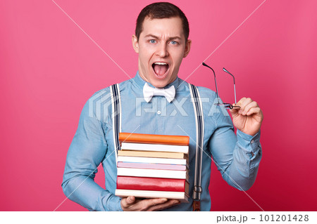 Handsome young man with stack of books looking at camera and screaming while standing on rosy background. Tired student finds out about additional tasks, prepares for classes. Education concept. 101201428