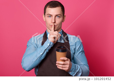 Dont tell secret of making coffee. Mysterious handsome male barista works in cafe, makes hush gesture, keeps fore finger over mouth, holds disposable cup of hot beverage, dressed in apron poses indoor Dont tell secret of making coffee. Mysterious handsome male barista works in cafe, makes hush gesture, keeps fore finger over mouth, holds disposable cup of hot beverage, dressed in apron poses indoor 101201452