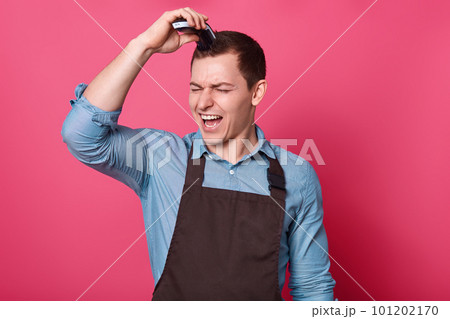 Positive man barber cuts hair with electric razor, makes trendy hairstyle, exclaims with joy, wears elegant shirt and apron, poses against pink background. Men, grooming, hairstyling, beauty concept. 101202170