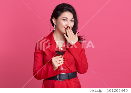 Close up portrait of brunette girl with glass of wine, wearing red dress with black belt, lady covering her mouth and looking directly at camera, female posing isolated over pink studio background. 101202449
