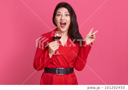 Horizontal shot of young celebrating woman red dress . Beautiful model posing isolated over pink background holding wine glass and showing v sign, screaming something, posing with widely opened mouth. 101202450