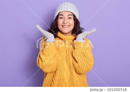 Beautiful woman with dark wavy hair wearing warm yellow sweater, mittens and hat, looking at camera with charming smile, spreading her palms aside, female posing isolated over lilac background. Beautiful woman with dark wavy hair wearing warm yellow sweater, mittens and hat, looking at camera with charming smile, spreading her palms aside, female posing isolated over lilac background. 101202516