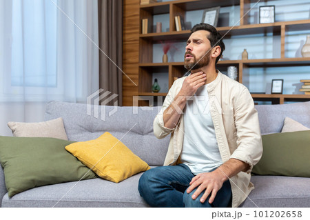 Sore throat. A young man sits at home on the sofa and holds his throat, feels a sore throat, burning sensation, discomfort. Sore throat. A young man sits at home on the sofa and holds his throat, feels a sore throat, burning sensation, discomfort. 101202658