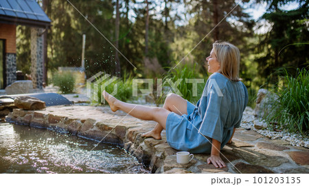 Happy young woman sitting by pond near cottege and enjoying cup of morning coffee on summer vacation in mountains. High angle view. 101203135