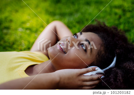 Multiracial girl lying down in grass and enjoying music in headphones, side view. Multiracial girl lying down in grass and enjoying music in headphones, side view. 101203182