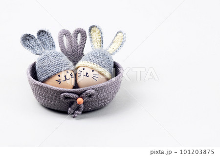 Two Easter eggs with funny faces and gray crocheted hats with bunny ears in a decorative basket on a gray background. Happy Easter concept. Greeting card. Two Easter eggs with funny faces and gray crocheted hats with bunny ears in a decorative basket on a gray background. Happy Easter concept. Greeting card. 101203875