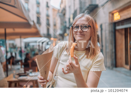 Happy woman eating traditional spanish delicious churros, a fried pastry with chocolate near a city cafe in Valence, Spain 101204074