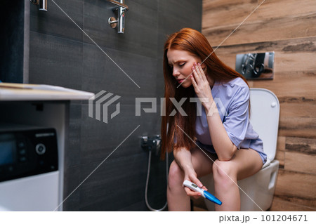 Portrait of frustrated young woman with anxious feelings holding negative pregnancy test sitting on toilet at home restroom. Unhappy redhead female holding pregnancy test feeling hopeless at bathroom. 101204671