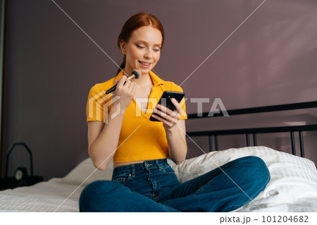 Vertical portrait of smiling redhead young woman applying cosmetic powder with big professional makeup brush and using smartphone. Pretty female using blusher with brush and reading social media. Vertical portrait of smiling redhead young woman applying cosmetic powder with big professional makeup brush and using smartphone. Pretty female using blusher with brush and reading social media. 101204682