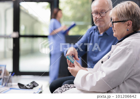 Senior medic taking patient insulin level test using glucometer in hospital waiting area, doing diabetes examination to measure glucose. Elderly man having checkup visit consultation 101206642