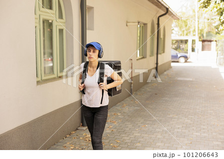 Food courier in headphones walking in street, standing outdoors, front view. Delivery by foot, woman going with customer order in thermal backpack, listening to music, delivering eatery meal 101206643