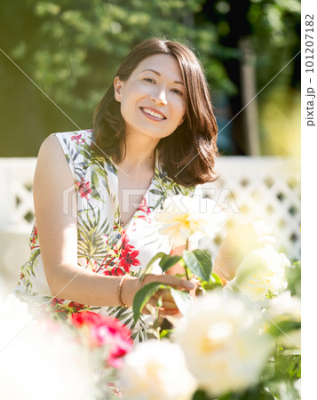 Wide smiling woman is admiring of blooming roses in public park. Summer vibes. Tropical plants and flowers in bloom in garden. Wide smiling woman is admiring of blooming roses in public park. Summer vibes. Tropical plants and flowers in bloom in garden. 101207182