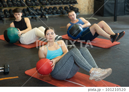 Strong young woman performing exercise for abs core with medicine ball on yoga mat during group workout in CrossFit gym center Strong young woman performing exercise for abs core with medicine ball on yoga mat during group workout in CrossFit gym center 101210520