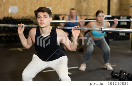 Motivated young man practicing exercises with rod near other people in gym Motivated young man practicing exercises with rod near other people in gym 101210882