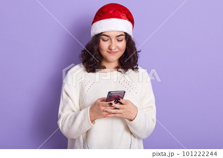 Image of attractive good looking young woman holding her smartphone, writing congratulations for New Year, wearing white sweater and red santa claus hat, isolated over lilac background in studio. 101222244