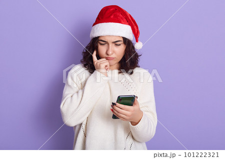 Thinking young woman in christmas santa hat and white knitted pullover, lady holding smart phone in hand, looking ar its screen with pensive expression, keeping finger on cheek. 101222321