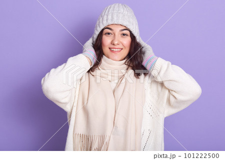 Horizontal portrait of attractive magnetic brunette putting grey hat on, wearing white sweater, scarf and gloves, looking directly at camera, standing isolated over lilac background in studio. 101222500