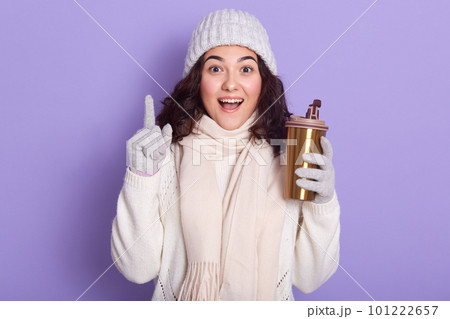 Image of surprised beautiful girl with black curly hair opening her mouth widely with shock, wearing light colored hat and gloves, white sweater and scarf, raising one finger, holding thermal cup. Image of surprised beautiful girl with black curly hair opening her mouth widely with shock, wearing light colored hat and gloves, white sweater and scarf, raising one finger, holding thermal cup. 101222657