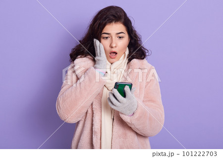 European woman touches cheek, being in shock, gets bill receipt for paying on mail, reads shocking news news from smart phone, has dark hair, wears rose faux fur coat coat, poses against lilac wall. European woman touches cheek, being in shock, gets bill receipt for paying on mail, reads shocking news news from smart phone, has dark hair, wears rose faux fur coat coat, poses against lilac wall. 101222703