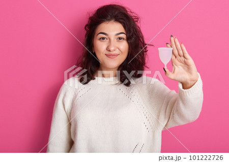 Image of confident charming brunette standing isolated over pink background in studio, holding mentrual cup in one hand, looking directly at camera, wearing white sweater. Menstruation concept. 101222726
