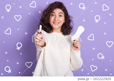Portrait of cheerful good looking female holding tampon on foreground, having hygiene pad in one hand, looking directly at camera, wearing white sweater, making choice. Critical days concept. Portrait of cheerful good looking female holding tampon on foreground, having hygiene pad in one hand, looking directly at camera, wearing white sweater, making choice. Critical days concept. 101222920