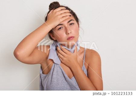 Woman puts hands on head and on throat, having scarf around neck, has high tempereature, lady wearing gray t shirt, posing isolated over white studio background. Healthy and illness concept. 101223133