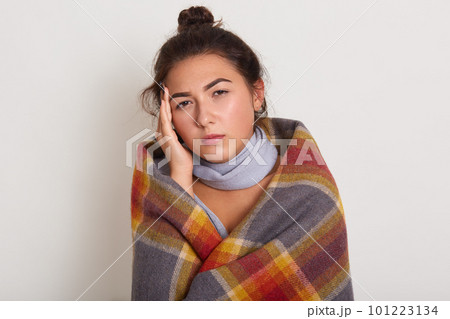 Sickness cute young woman with dark hair, wrapped warm checkeredplaid with headache, touching her head, keeps hand on temple, feeling first symptoms of illness. Flu, feeling unwell, cold concept 101223134