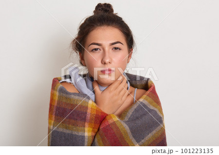 Image of sick young dark haired woman with bun on head, posing isolated over white studio background, unhealthy lady touches neck, wearing checkered plaid, lady has sore throat or catch cold. 101223135