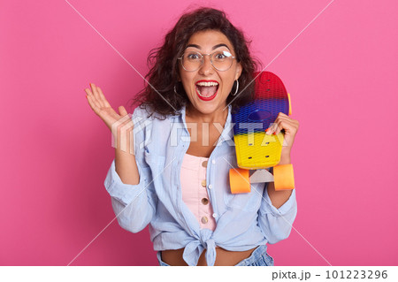 Horizontal shot of surprised young woman shouting over pink background, looking at directly camera with astonished and surprised facial expression, holding skateboard on her shoulders. People emotions Horizontal shot of surprised young woman shouting over pink background, looking at directly camera with astonished and surprised facial expression, holding skateboard on her shoulders. People emotions 101223296
