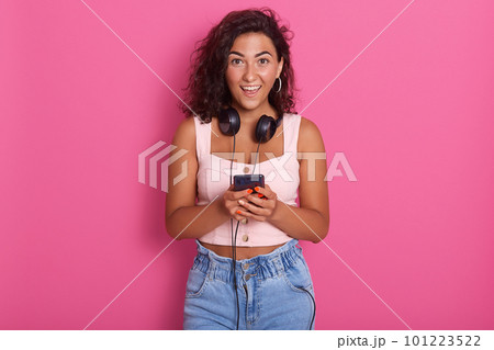 Portrait of happy beautiful young woman holding phone in hand and smiling, wearing casual clothing, posing isolated over pink background, female with dark wavy hair, expressing astonishment. Portrait of happy beautiful young woman holding phone in hand and smiling, wearing casual clothing, posing isolated over pink background, female with dark wavy hair, expressing astonishment. 101223522