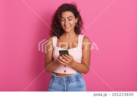 Woman with mobile phone in hands, cheerful young woman holding smart phone and smiling while standing isolated over pink backgrond, female wearing rosy top and jeans,checking social network. 101223830