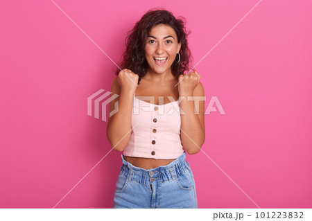 Photo of happy young woman with beautiful dark wawy hair screaming and clenching fist while looking directlyat camera, wearing stylish outfit, isolated over pink background. People emotions concept. 101223832