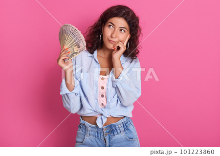 Young attractive smiling woman thinking how to spend her bunch of money, female wearing blue shirt and jeans, standing against pink wall with pensive expression. Young attractive smiling woman thinking how to spend her bunch of money, female wearing blue shirt and jeans, standing against pink wall with pensive expression. 101223860