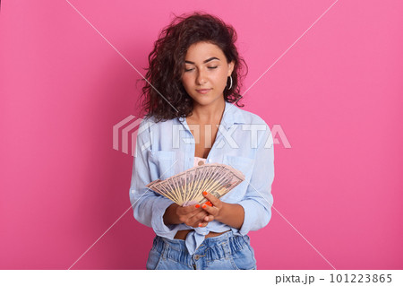 Image of thounghtful young brunette woman wearing blue shot and jeans standing isolated over pink studio background. Looking at fan of money in her hands, counting her salary. People concept. Image of thounghtful young brunette woman wearing blue shot and jeans standing isolated over pink studio background. Looking at fan of money in her hands, counting her salary. People concept. 101223865