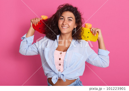Image of cheerful good looking female with dark curly hair smiling sincerely, having pleasant facial expression, holding her colorful skateboard in hands aback, being in high spirits. Mood concept. Image of cheerful good looking female with dark curly hair smiling sincerely, having pleasant facial expression, holding her colorful skateboard in hands aback, being in high spirits. Mood concept. 101223901