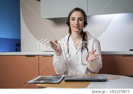 Young female doctor in a white medical uniform with a stethoscope, using a computer laptop, talks via videoconference with a patient, looking into the camera at a healthcare hospital 101223977
