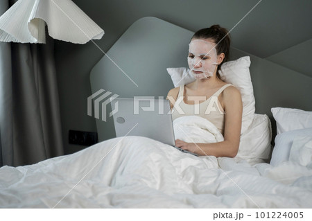 Healthy woman is sitting on a bed with a cosmetic face mask, using a laptop and working online. Have a cozy time at home. The concept of self-service and home spa. 101224005