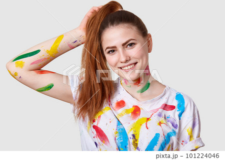 Headshot of pleased cheerful woman has dark hair combed in pony tail, smiles gently, has dirty body and t shirt with acrylic paints, brings creativity to life, isolated over white studio wall Headshot of pleased cheerful woman has dark hair combed in pony tail, smiles gently, has dirty body and t shirt with acrylic paints, brings creativity to life, isolated over white studio wall 101224046