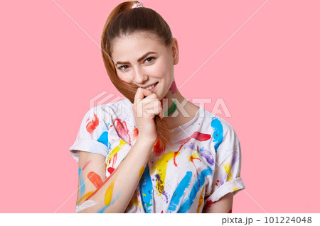 Isolated shot of pleased artistic woman with pony tail, has stained t shirt with watercolours, has creatie occupation, enjoys hobby, poses against pink background. Process of creation. Art concept 101224048