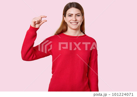 Horizontal shot of attractive young woman shows tiny object, gestures with hands, dressed in red clothes, has toothy smile, demonstrates how much she recieves money, models over rosy background 101224071