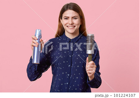 Indoor shot of happy young woman with dark hair holds hairspray and comb, dressed in blue shirt, smiles broadly, isolated over rosy bacground, makes coiffure. People and positiveness concept 101224107