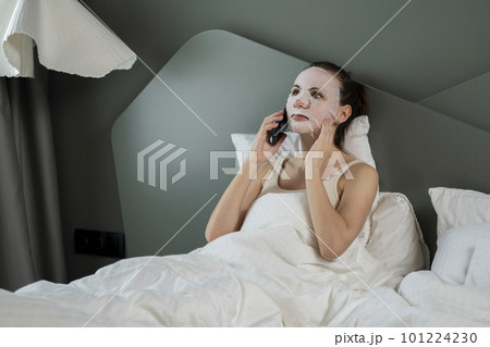 Woman with a cosmetic face mask is sitting on the bed, talking on the phone at home in the bedroom Woman with a cosmetic face mask is sitting on the bed, talking on the phone at home in the bedroom 101224230