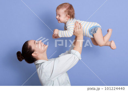 Portrait of happy mother and child together having fun isolated over blue background, attractive female with bun, dresses white shirt, infant wearing striped bodysuit. Mom and son, family concept. Portrait of happy mother and child together having fun isolated over blue background, attractive female with bun, dresses white shirt, infant wearing striped bodysuit. Mom and son, family concept. 101224440