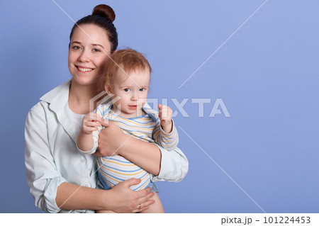 Horizontal shot of happy smiling mum holding her little son in hands, infant wearing striped bodysuit showing his fists, poses isolated on blue background. Copy space for advertisement or promotion Horizontal shot of happy smiling mum holding her little son in hands, infant wearing striped bodysuit showing his fists, poses isolated on blue background. Copy space for advertisement or promotion 101224453