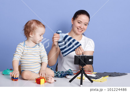 Close up portrait of contemporary young woman holding knitted cap while filming video for fashion and beauty channel against blue background, posing with her child isolated over blue background. 101224486