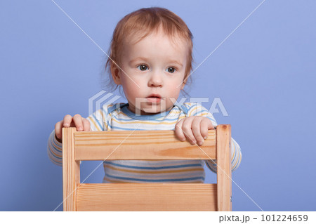 Close up portrait of little child looking aside, standing on chair isolated over blue background in studio, wearing striped sweater, being attentive, focused on something. Childhood concept. 101224659