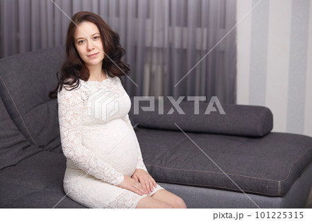 Beautiful young future mom in gentle white dress, has dark wavy hair, sits on grey couch sofa at home, looks at camera, being photograpfed for album of memories about pragnancy. Motherhood concept. 101225315