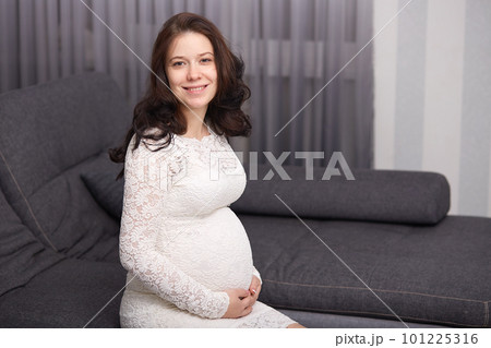 Young smiling happy pregnant lady sits on sofa in cosy living room, looks directly at camera, wearing formal white dress, having beautiful hairdo, holding both her hands on belly, looks happy. 101225316