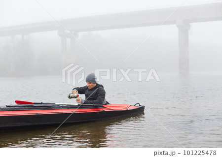 handsome young male in canoe in middle of river, pouring hot beverage from thermos, guy wearing black jacket and gray cap enjoying coffee or tea in kayak in open air on beautiful foggy autumn day. handsome young male in canoe in middle of river, pouring hot beverage from thermos, guy wearing black jacket and gray cap enjoying coffee or tea in kayak in open air on beautiful foggy autumn day. 101225478
