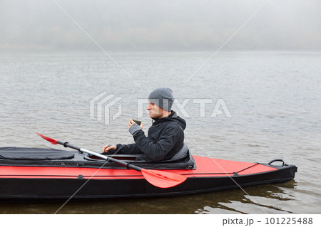 Man paddles red and black kayak and sitting with paddle, holds mug with hot coffee or tea on river or lake in fall season. Autumn kayaking, water extreme sport. 101225488